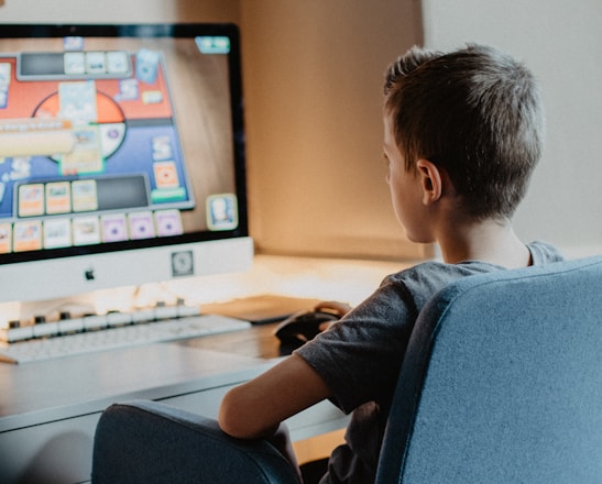 A young person is sitting on a blue chair at a desk, focused on a computer screen displaying a colorful card game. The scene appears to be set in a cozy, well-lit room.