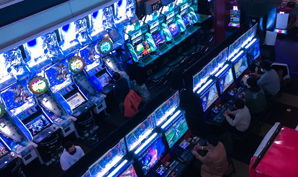 A bustling arcade filled with rows of gaming machines, each with vibrant displays and controls. Several people are seated at these machines, deeply engaged in playing games. Bright fluorescent lights cast a blue and neon glow across the room, enhancing the lively and energetic atmosphere.